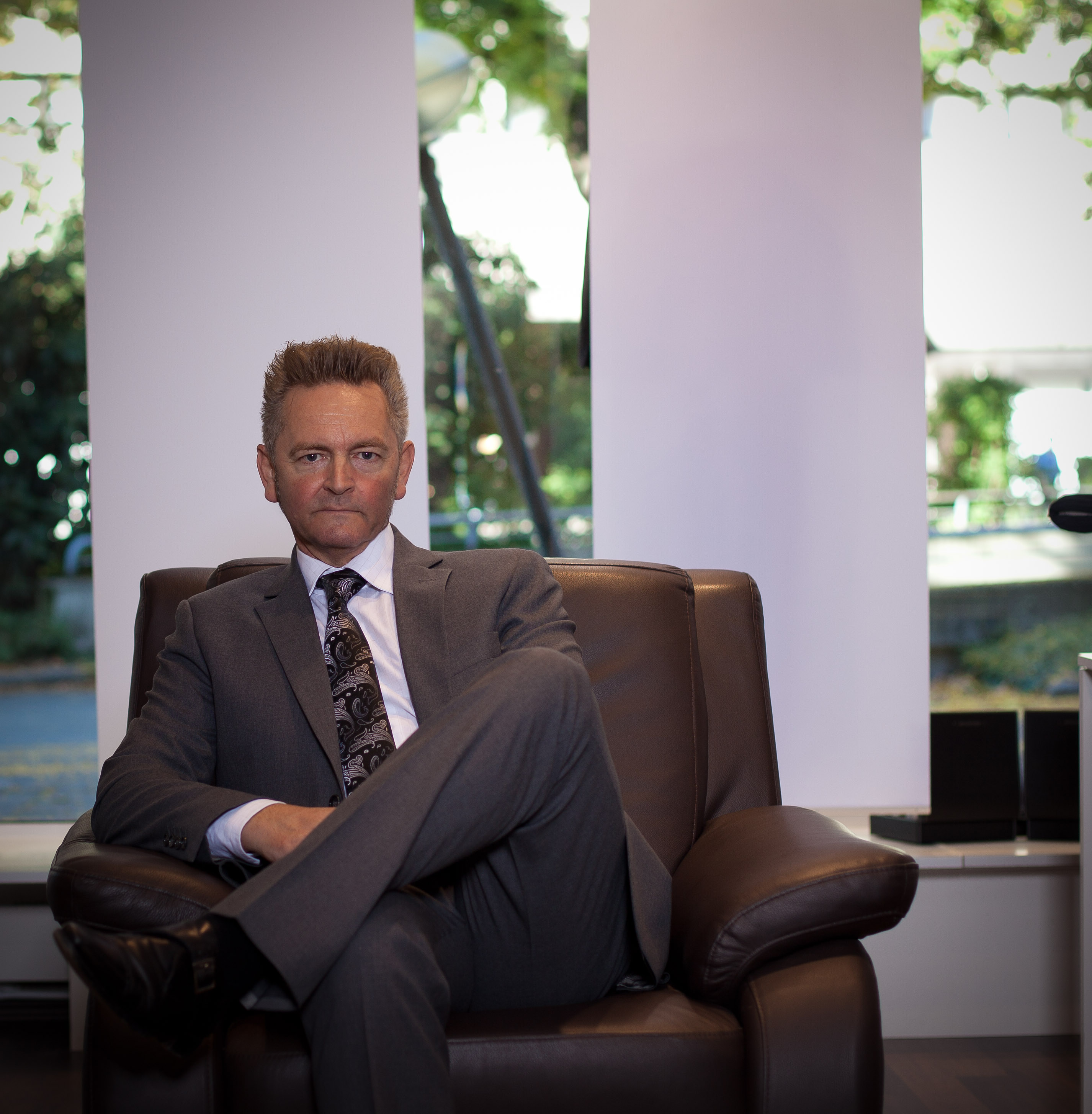 Ein eleganter Geschäftsmann in einem perfekt sitzenden, maßgeschneiderten Anzug sitzt entspannt auf einem braunen Ledersessel in einem modern gestalteten Showroom. Der Anzug besticht durch eine exzellente Passform und hochwertige Stoffe, ergänzt durch eine stilvolle Krawatte mit dezentem Muster. Im Hintergrund sorgen große Fenster für natürliches Licht und bieten einen Blick ins Grüne, was dem Raum eine helle und einladende Atmosphäre verleiht. Dieses Bild symbolisiert die Professionalität und den exklusiven Service von Baran Maßkonfektion, Ihrem Experten für individuelle Maßanzüge in Walldürn.