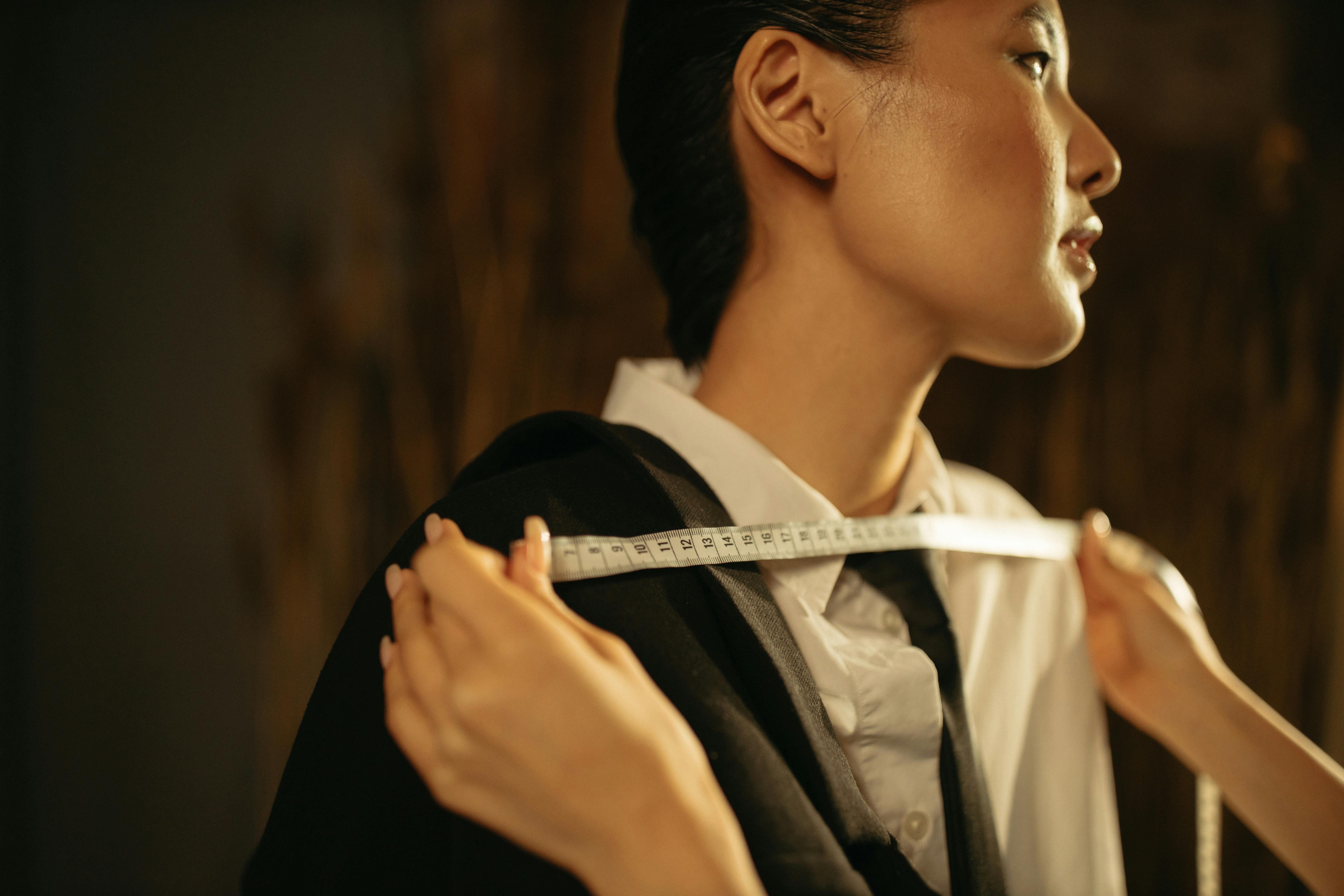 Eine Schneiderin misst sorgfältig die Schulterbreite einer Kundin für einen maßgeschneiderten Anzug. Die Frau trägt ein weißes Hemd mit schwarzer Krawatte, während ein Maßband präzise über ihre Schultern gelegt wird. Das Bild vermittelt Professionalität und Handwerkskunst, die für individuelle Maßanfertigungen unerlässlich sind. Die Szene hebt die Detailgenauigkeit und den persönlichen Service hervor, den Baran Maßkonfektion bei der Anfertigung von perfekt sitzender Kleidung für Damen und Herren bietet